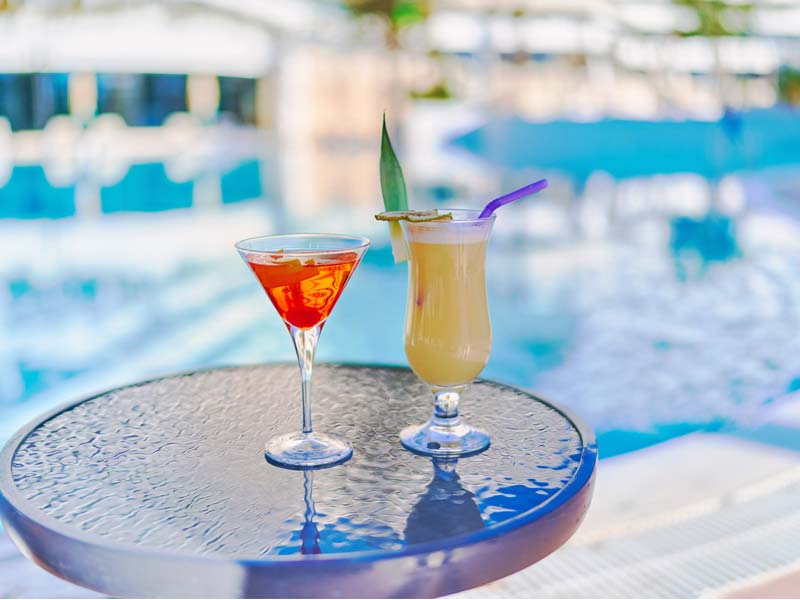 Two exotic cocktails on a tray, with a backdrop of a hotel swimming pool. 